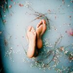 Bare feet submerged in pastel water adorned with delicate petals and flowers, creating a tranquil scene.